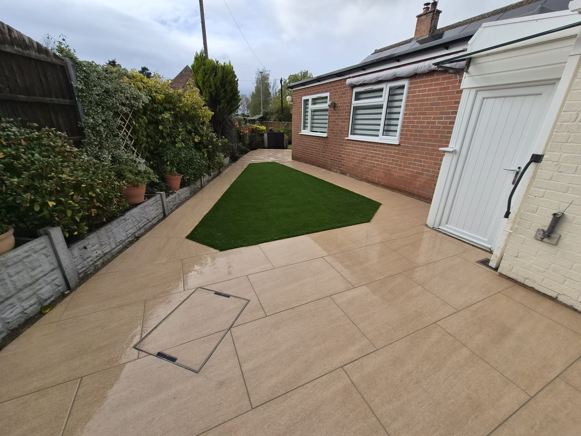 Artificial grass lawn surrounded by beige paving beside a brick bungalow